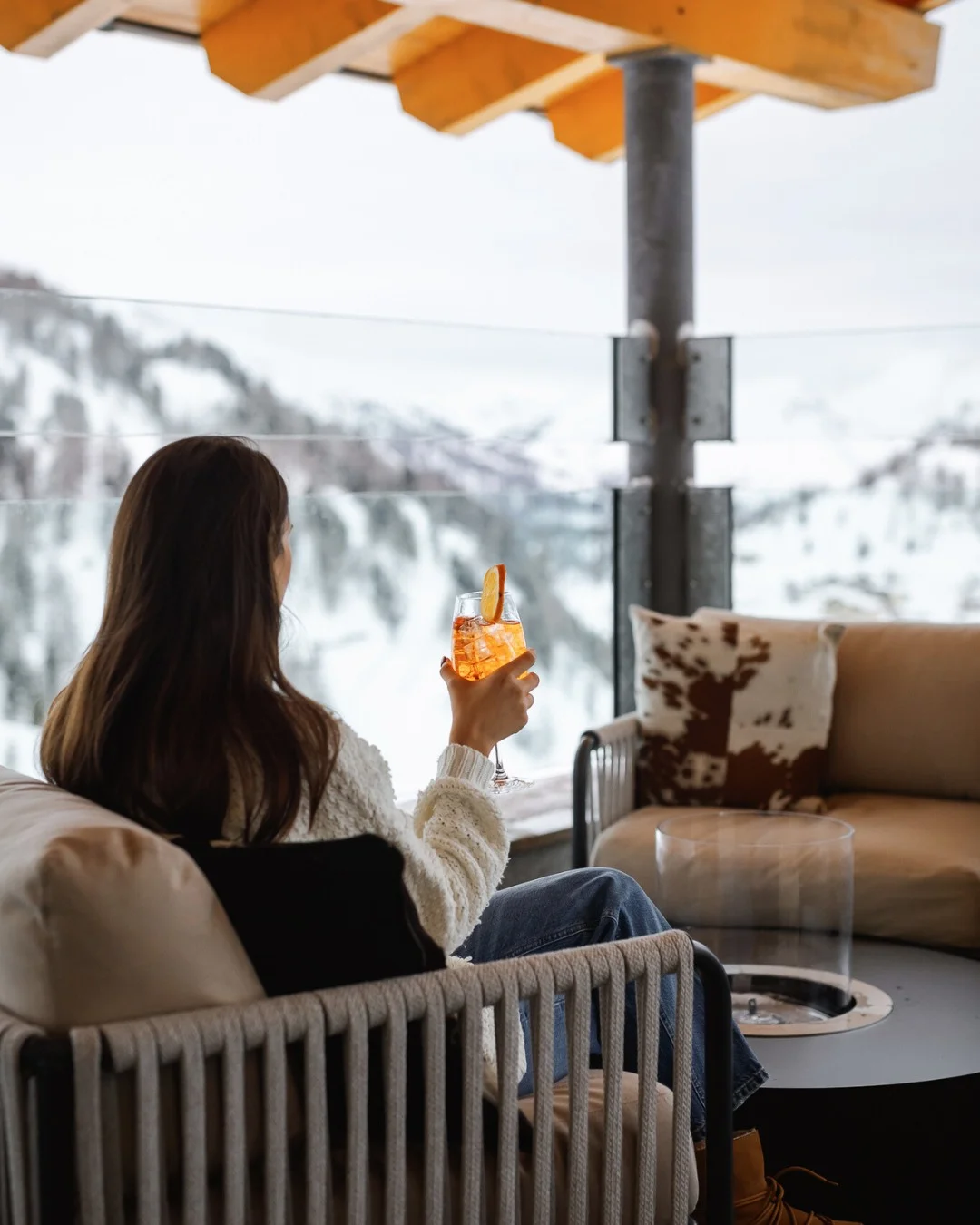 A woman sitting on a rooftop terrace overlooking snow-covered mountains, holding a glass of Aperol Spritz.