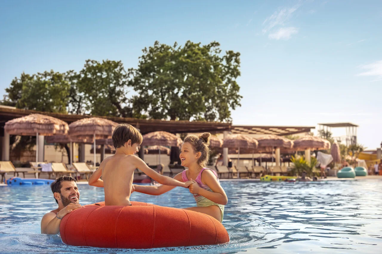 A father and two children play in a pool on a large orange inflatable ring, with sun loungers and parasols in the background