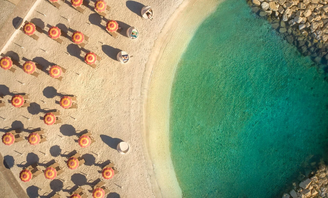 Pogled odozgo na pješčanu plažu Maro Sandy u Istri s pravilno raspoređenim crveno-žutim suncobranima, ležaljkama i čistim morem.