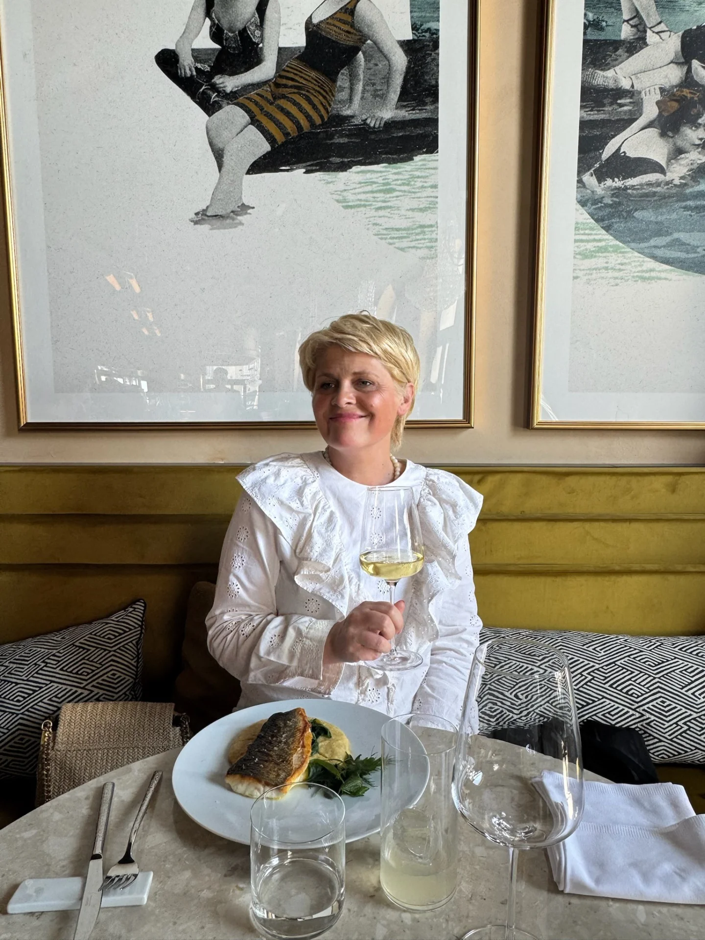 A woman seated in a restaurant holding a glass of white wine, with a seafood dish on the table.