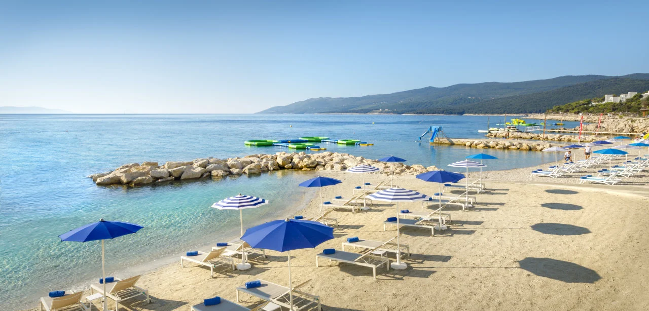 Prostrana pješčana plaža u Rapcu s plavim suncobranima, bijelim ležaljkama i morskim parkom na napuhavanje u iznimno bistroj uvali.