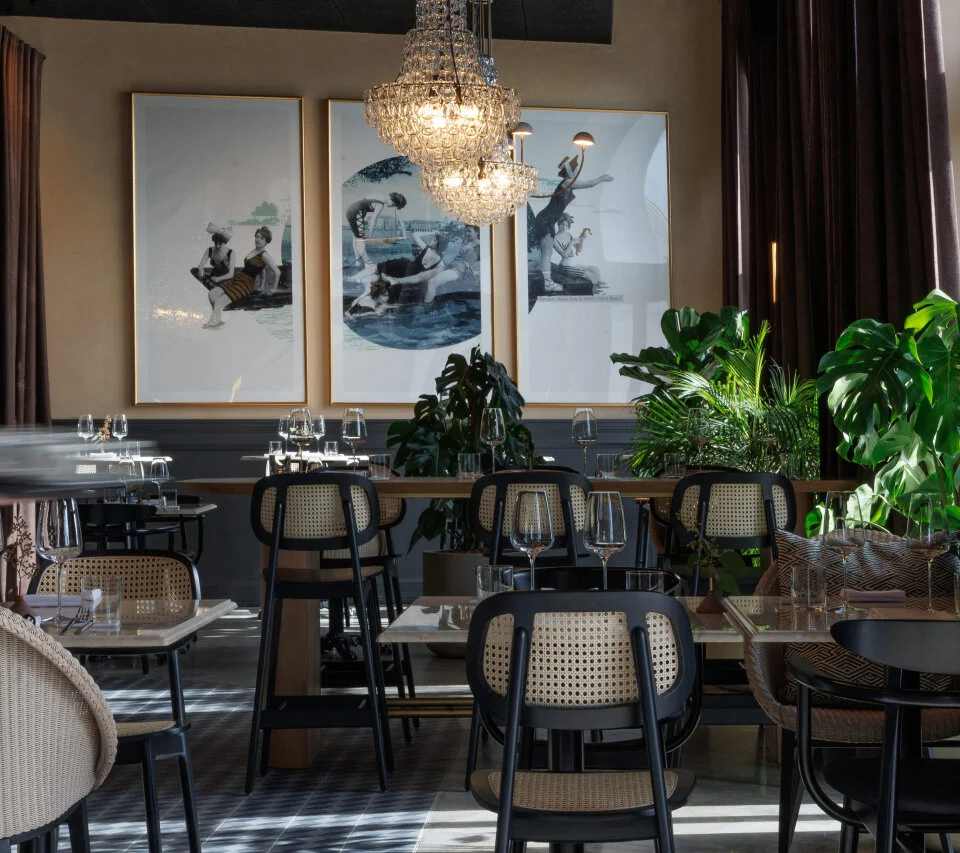Elegant restaurant interior with rattan chairs, framed artwork, plants, and a crystal chandelier.