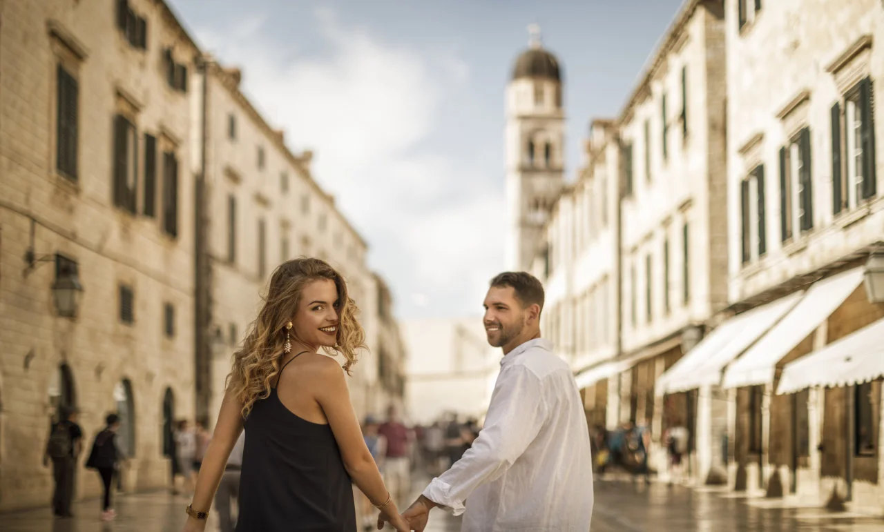 A couple walks hand in hand along a historic street in Dubrovnik's Old Town, with the ancient city walls and bell tower in the background on a sunny day.