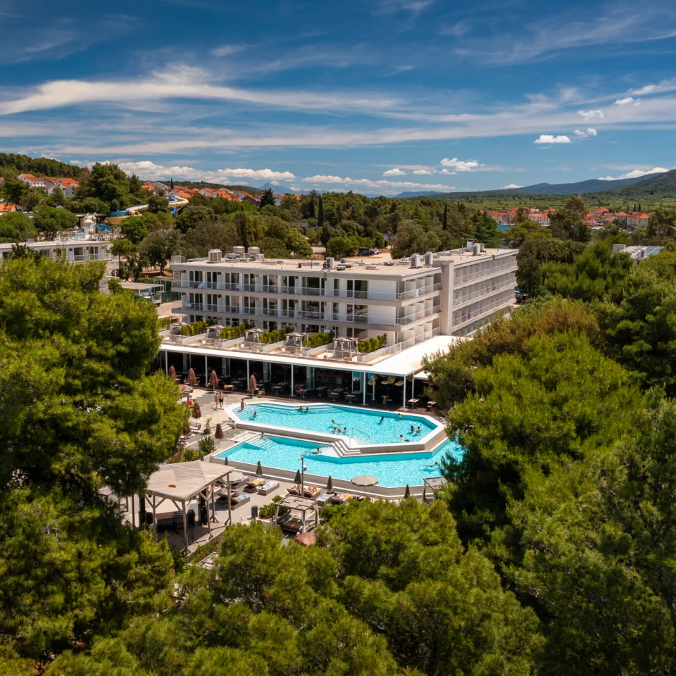 Exterior view of [PLACES] Hvar by Valamar hotel featuring an outdoor pool surrounded by pine trees.