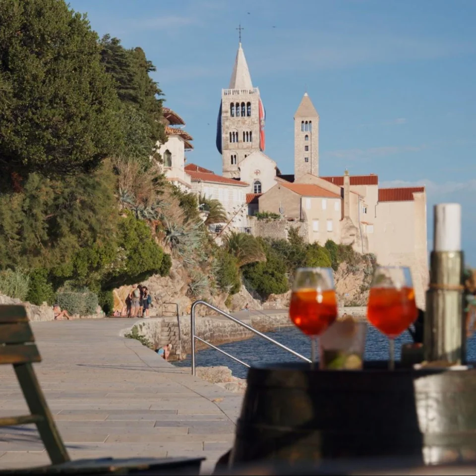 View from Banova Villa Beach Bar towards the sea and the bell towers of Rab Old Town, with two cocktails in the foreground.