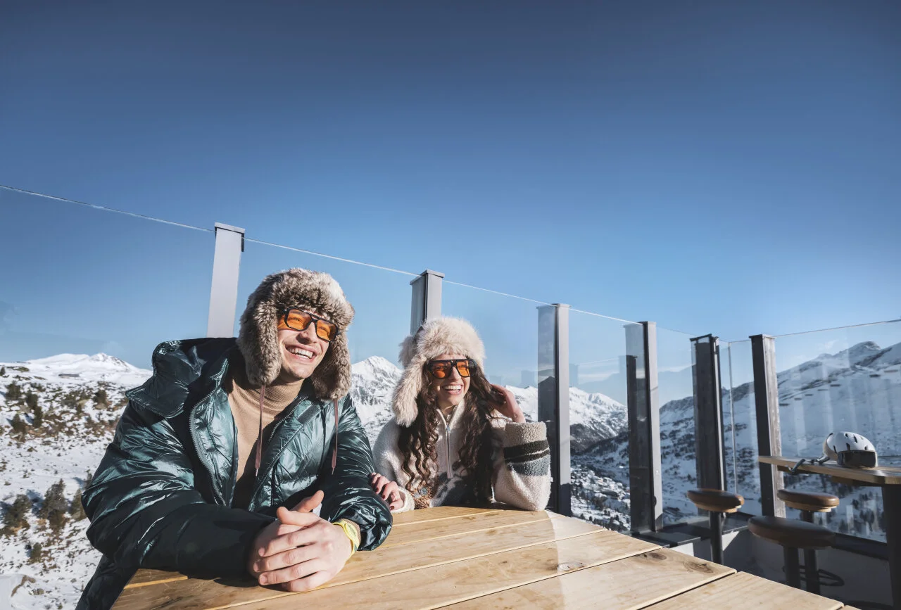 Two people enjoying a sunny winter day on a mountain terrace with panoramic views of snow-covered peaks.