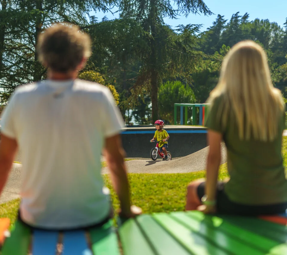 Parents watching their child ride a bike in a green outdoor play area.