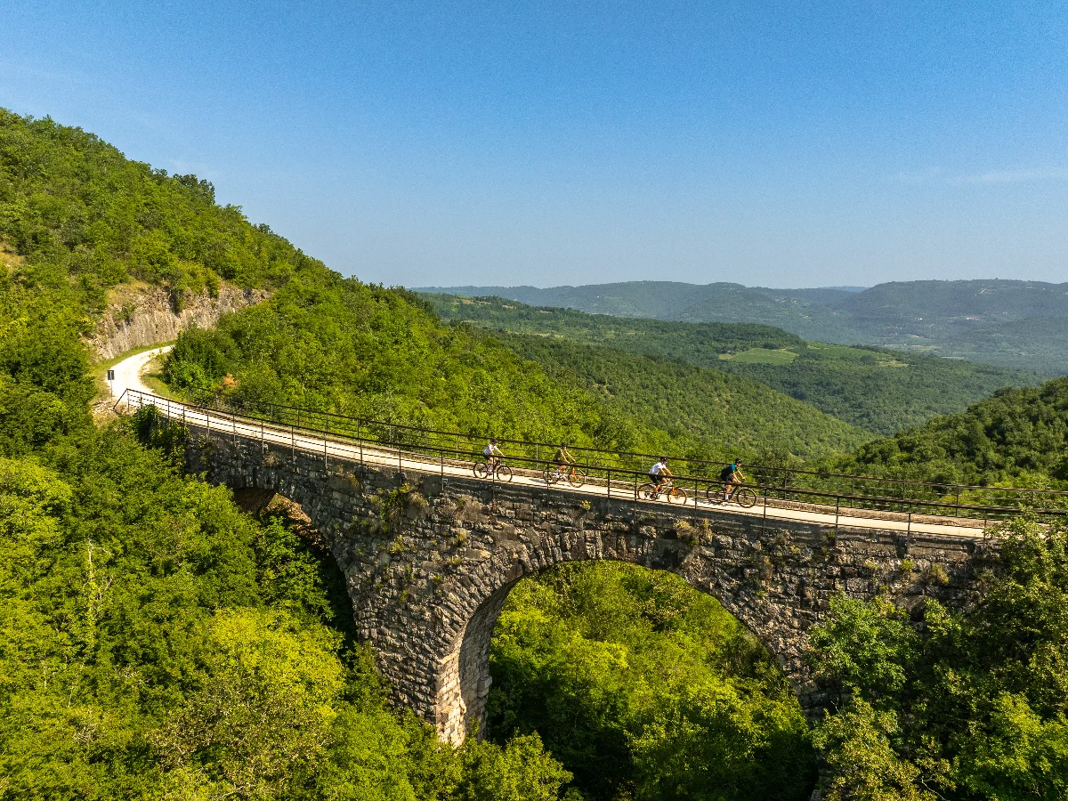 Grupa biciklista vozi Parenzanom i prelazi preko starog kamenog mosta, uz pogled na zelene istarske brežuljke.