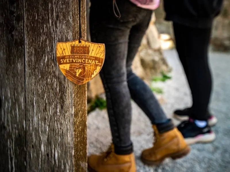 Drveni privjesak s natpisom “Escape Castle Svetvinčenat” visi na starim vratima, dok u pozadini posjetitelji istražuju prostor.