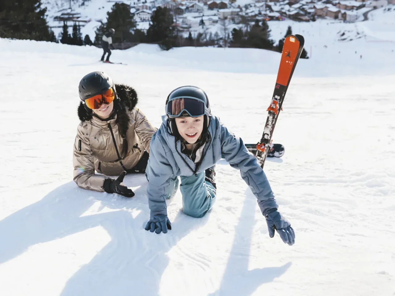 Mama i sin leže u snijegu na skijaškoj stazi, a u pozadini se vide snijegom prekrivene planine i skijalište.