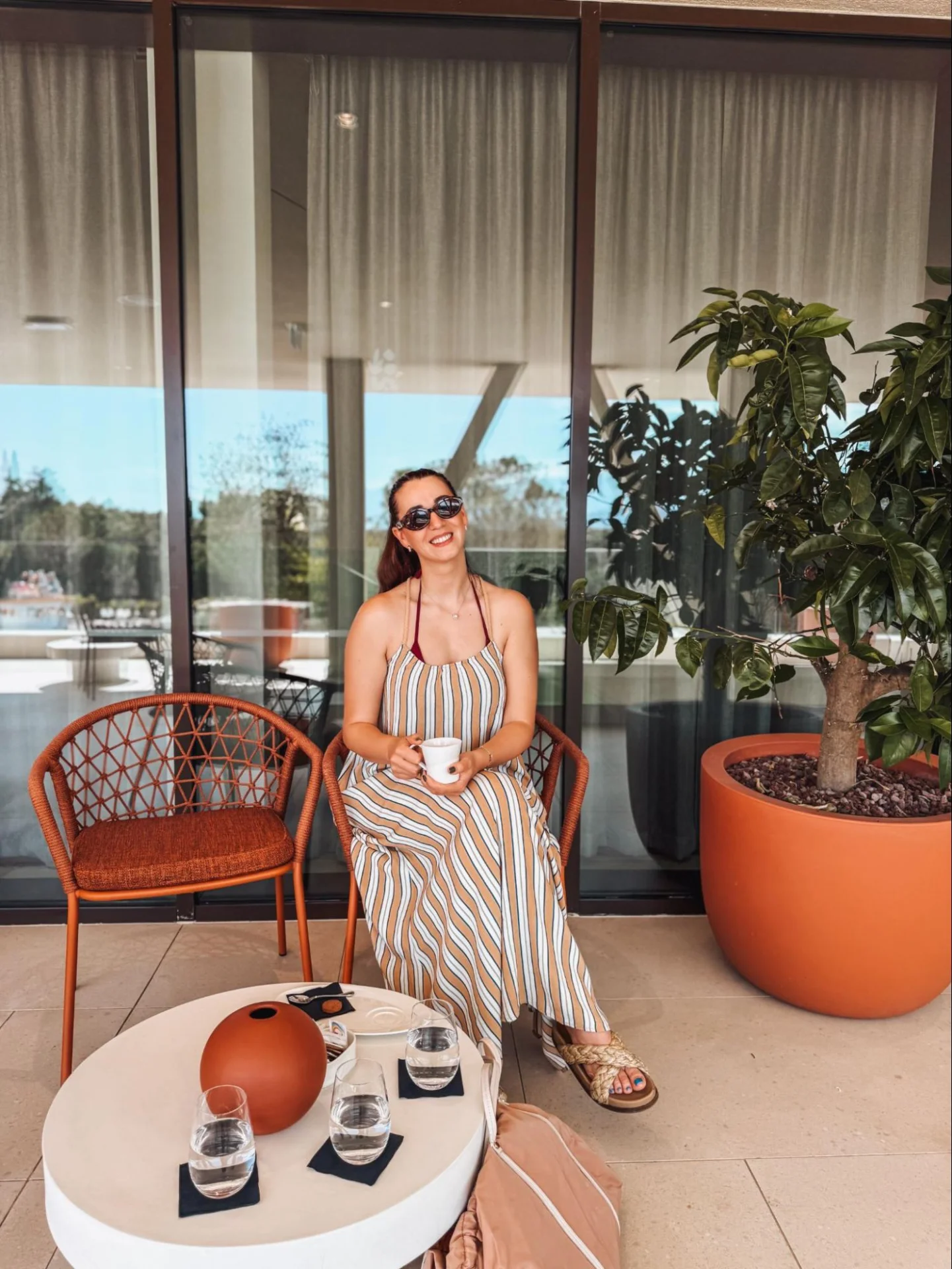 A woman in a striped dress is sitting on the terrace of Arba Resort at a table with coffee and serving details.