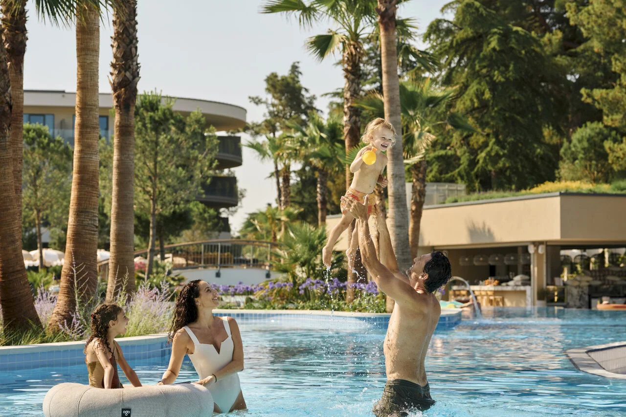 Family enjoying playtime in an outdoor swimming pool surrounded by greenery and palm trees.