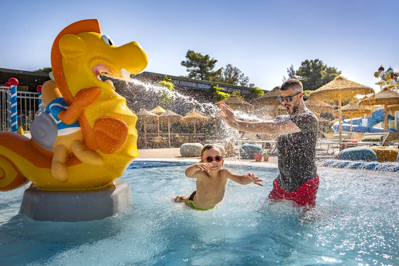Parent and child playing together in a children's pool with water attractions and slides.